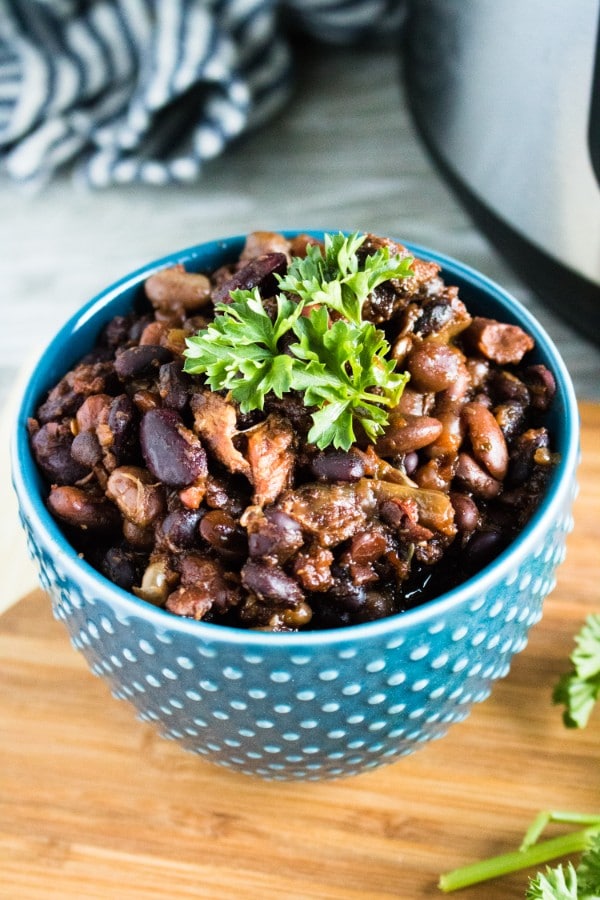 a bowl of pork and beans in a blue bowl topped with parsley on a cutting board with an instant pot in the background