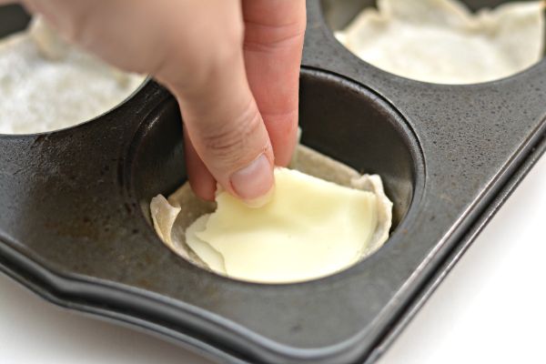 a hand placing provolone cheese on top of a wonton wrapper in a muffin tin