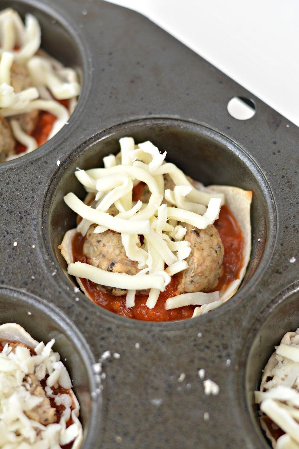 a muffin tin filled with wonton wrappers, sauce, mini meatballs, and shredded cheese