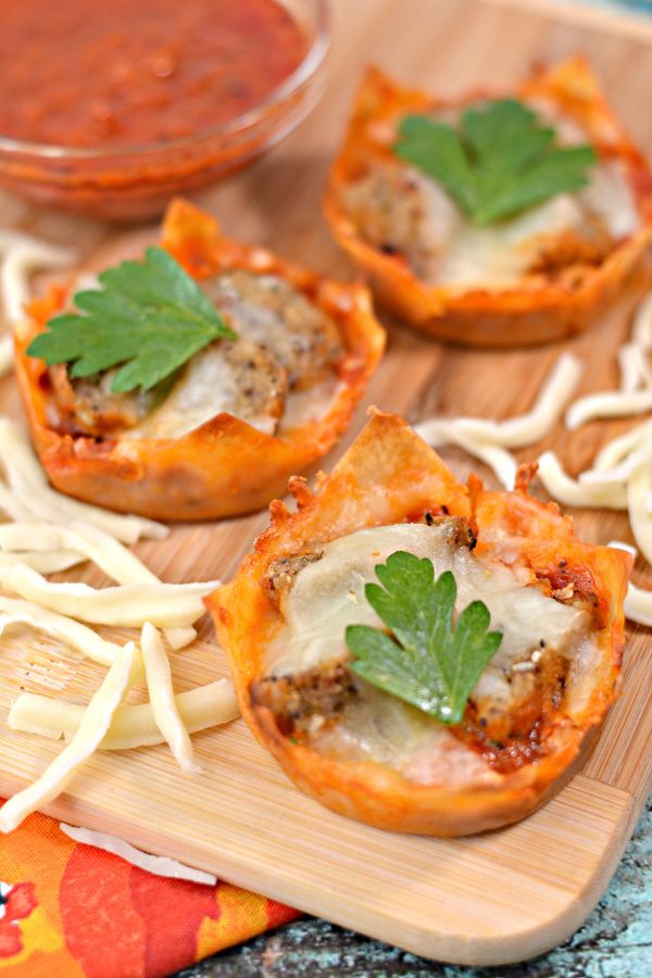 mini meatball appetizer cups on a cutting board next to shredded mozzarella cheese and a glass bowl of sauce