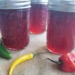 Three jars of red pepper jelly with metal lids are placed on a burlap surface, surrounded by whole green, yellow, and red chili peppers, perfect for those interested in a raspberry hot pepper jelly recipe.