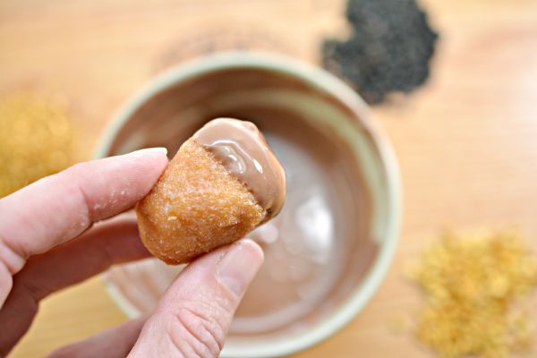 a hand holding a donut hole in the shape of an acorn dipped in chocolate with the bowl of chocolate in the background