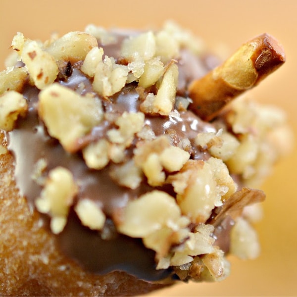 a closeup of a donut hole in the shape of an acorn dipped in chocolate and nuts with a pretzel stem