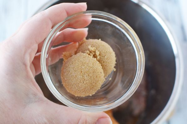 a lady's hand holding a glass bowl of brown sugar over an instant pot