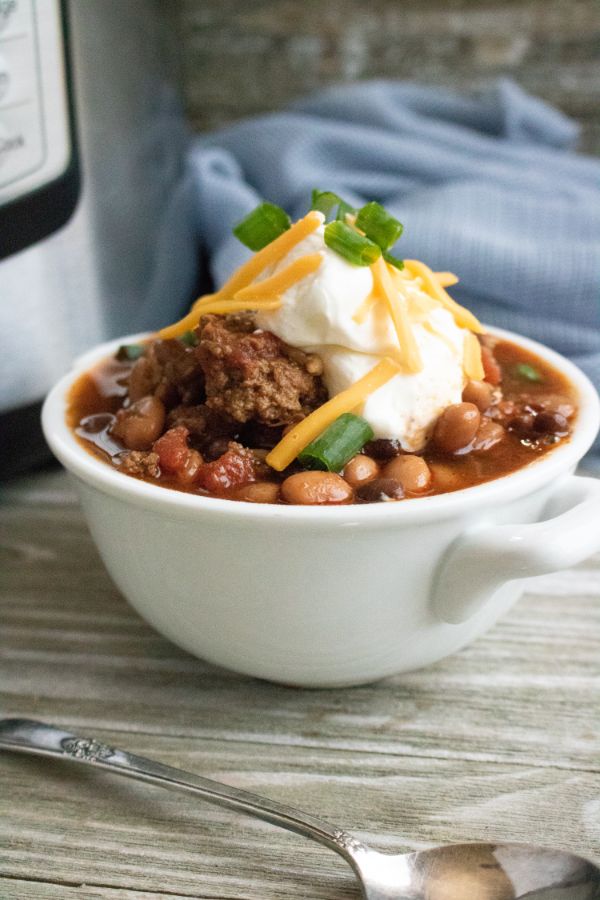 chili in a white bowl topped with sour cream, cheese, and green onions on a table next to a spoon and an instant pot