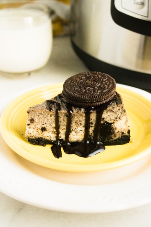 a slice of oreo cheesecake on a yellow and white plate topped with chocolate sauce and an oreo with a glass of milk and an instant pot in the background