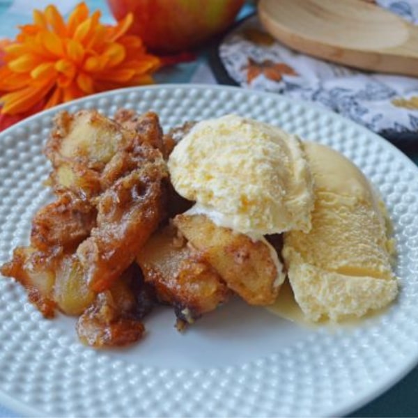 apple crisp on a plate topped with vanilla ice cream with and orange flower, apple, and wooden spoon in the background
