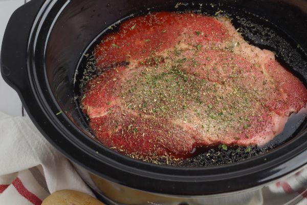 a crockpot with a beef roast in it topped with seasonings. with a red and white linen under it