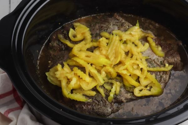 a crockpot with a roast in it, covered in sauce and peppers