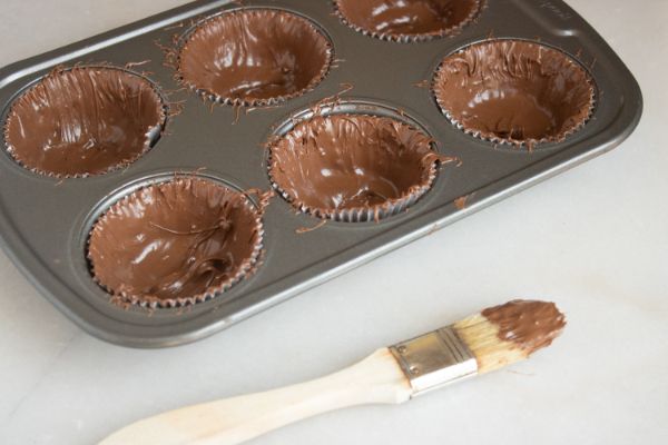 a pastry brush with chocolate on it next to a muffin tin filled with paper liners spread with chocolate