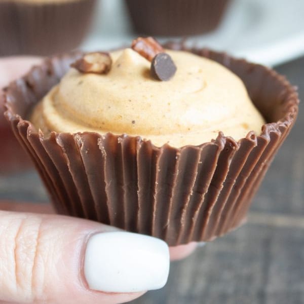 a lady's hand holding a chocolate cup filled with pumpkin mousse topped with nuts and a chocolate chip