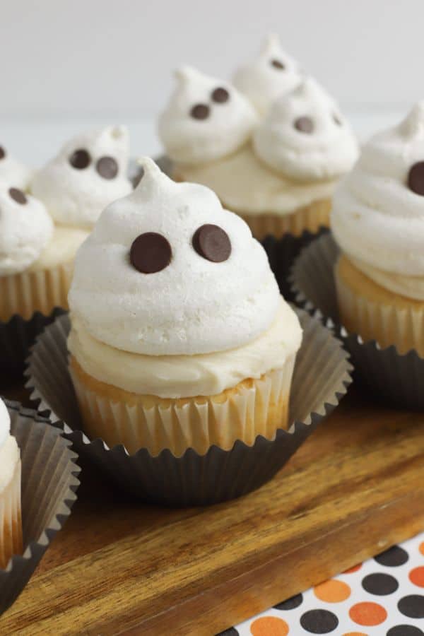 cupcakes topped with vanilla frosting and a meringue cookie with chocolate chips in it so it looks like a ghost with eyes, all on a wood slat