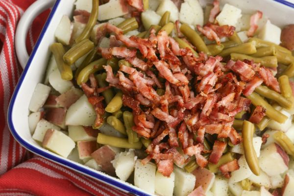potatoes in a white casserole dish topped with green beans and bacon, all on a red linen