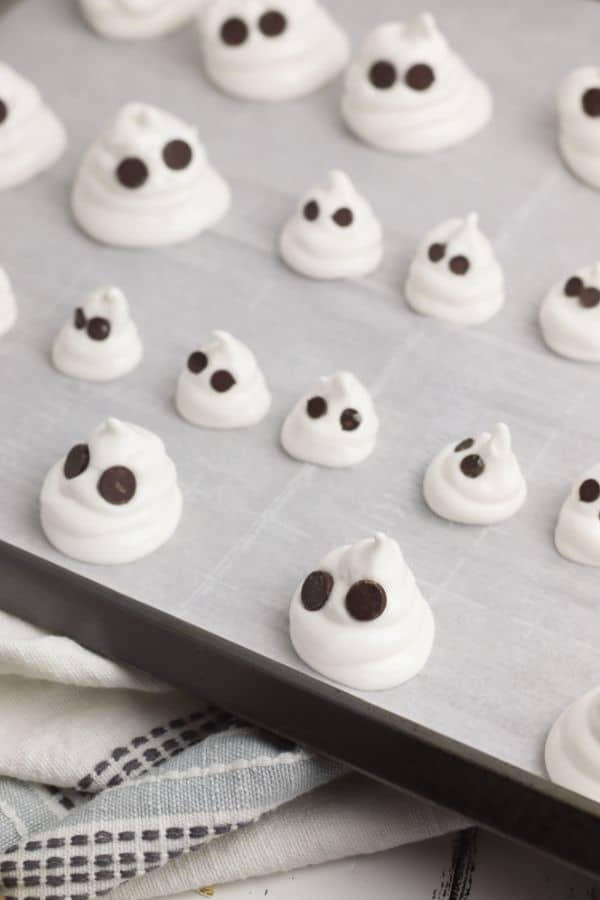 meringue batter piped onto a parchment paper on a baking sheet to look like ghosts with two chocolate chip eyes in the batter