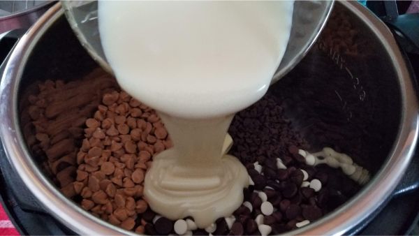 sweetened condensed milk being poured from a bowl into the Instant Pot that's filled with peanut butter chips, white chocolate chips, and chocolate chips