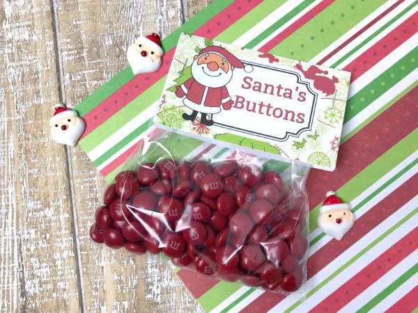 a plastic bag filled with red m&ms with a Santa's Buttons label on a red, green & white striped paper with mini Santa faces on it on a brown table