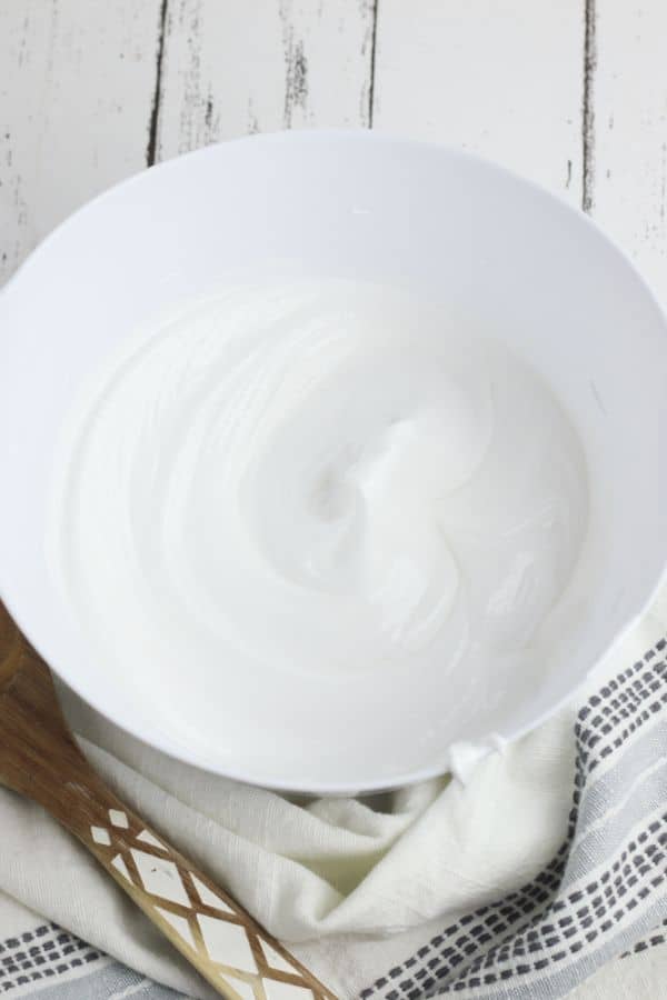 stiff egg whites in a white mixing bowl on a white wood table with a gray linen next to them