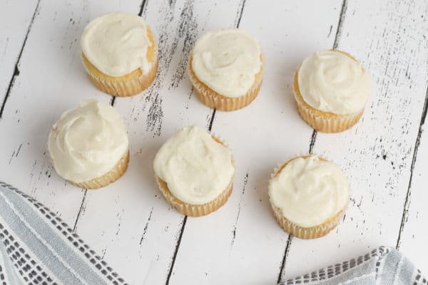 white cupcakes topped with vanilla frosting on a white wood table with a gray linen next to them