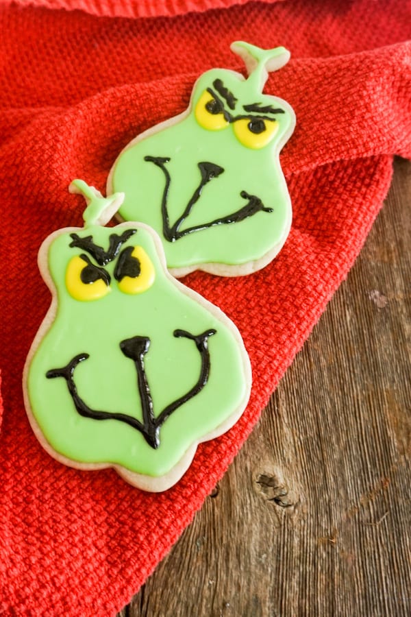 cookies in the shape of the Grinch, frosted with green, black and yellow frosting to look like the Grinch on a red linen on a wood table