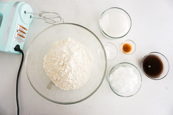 a hand mixer next to glass bowls of flour, sugar, baking powder, vanilla extract, aquafaba, coconut oil, on a white counter