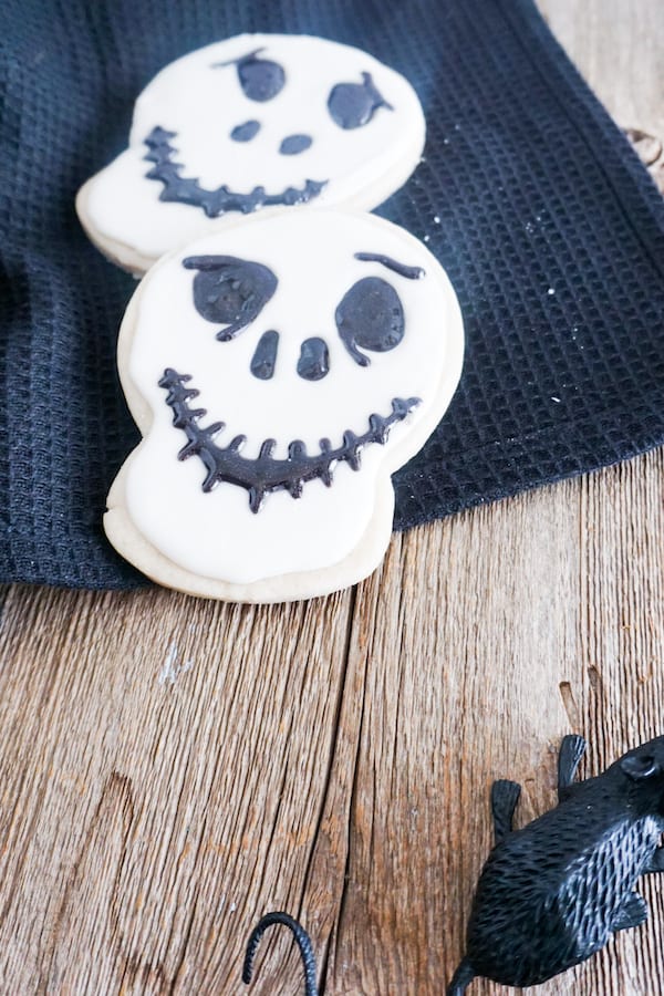 two cookies decorated with white frosting and black gel to look like Jack Skellington on a black linen on a wood table with a fake mouse next to it