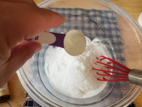 a hand pouring witch hazel into a glass bowl of baking soda and soap with a whisk in it