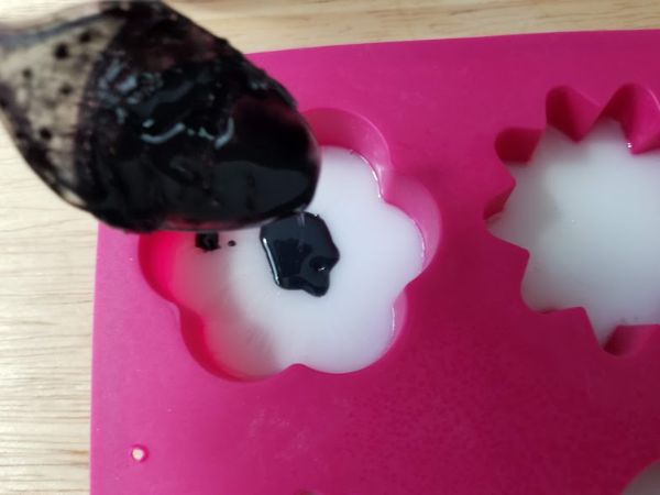 a spoon dropping black soap mixture onto white soap mixture into a soap mold on a wooden table