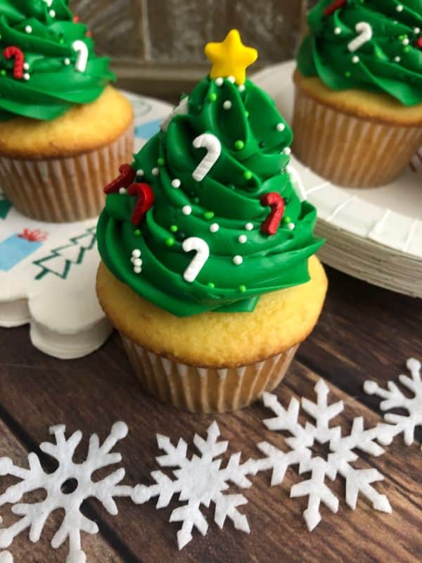 Several cupcakes with green frosting on top in the shape of a Christmas tree, with candy cane sprinkles and a star on top. Snowflakes are in the background.