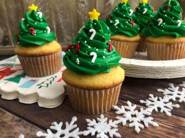 Several cupcakes with green frosting on top in the shape of a Christmas tree, with candy cane sprinkles and a star on top. Snowflakes are in the background.