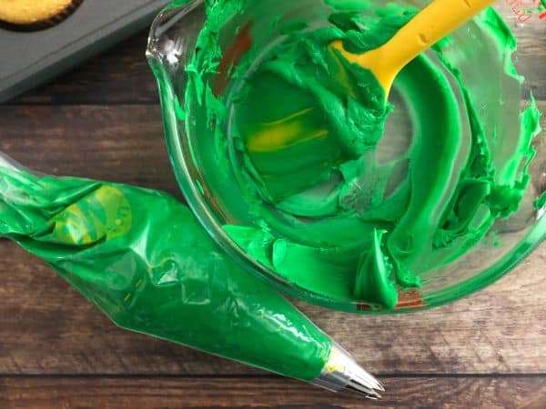 Green frosting in a glass bowl with a yellow spoon inside with a frosting bag next to it and a cupcake tin in the background.