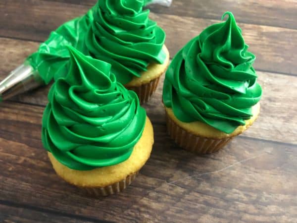 Three cupcakes with green frosting on top in the shape of a Christmas tree with a frosting bag in the background.