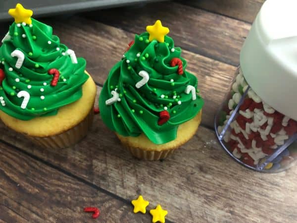Two green cupcakes with green frosting in the shape of a Christmas tree with candy cane sprinkles and a star on top, and sprinkles in a container in the background.