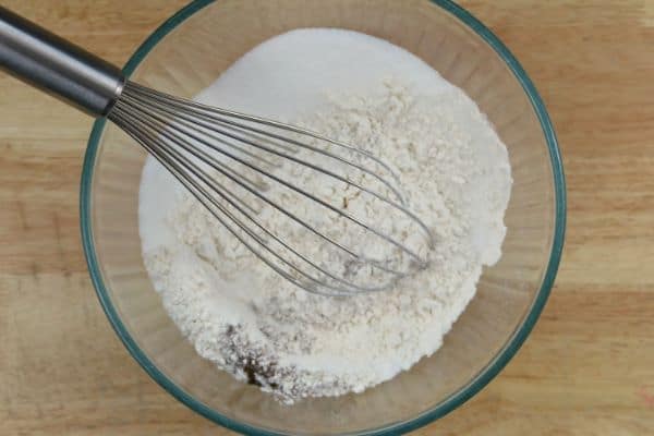 A glass bowl with flour, baking soda and other dry ingredients with a wire whisk in the bowl.