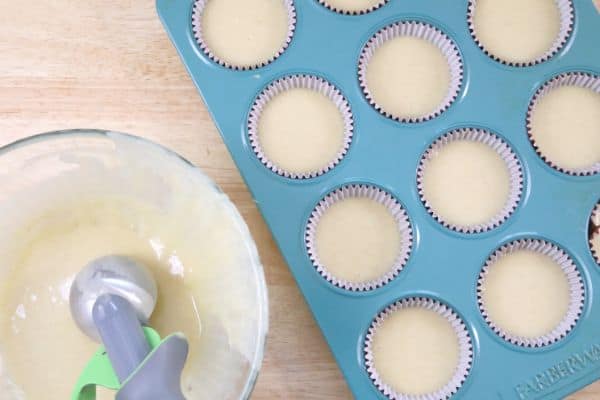 A cupcake pan full of cupcake liners filled with cupcake batter next to a glass bowl with cupcake batter in it and a cookie scoop