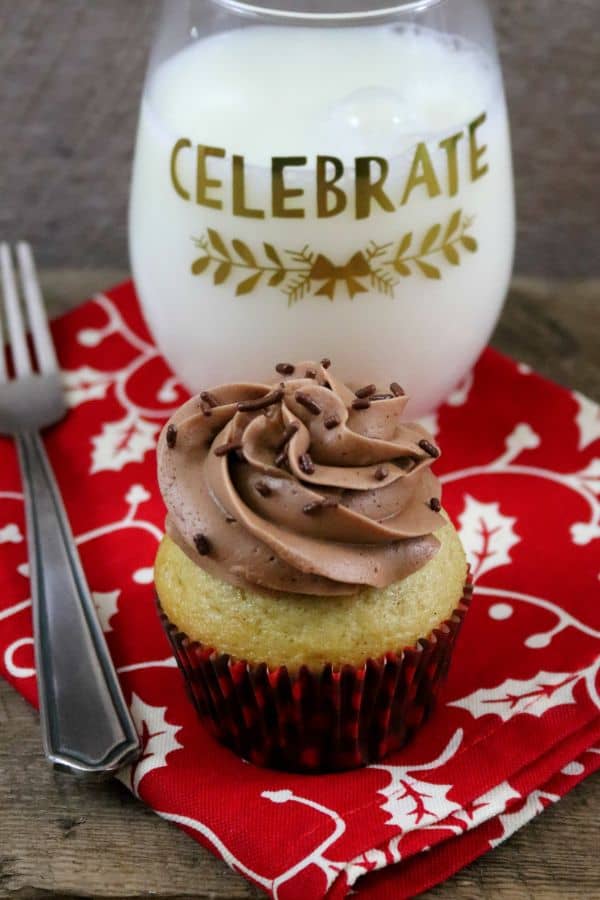 An eggnog cupcake with chocolate frosting and chocolate sprinkles on a red and white napkin with a fork next to it and a glass behind it filled with milk with the words celebrate on it.
