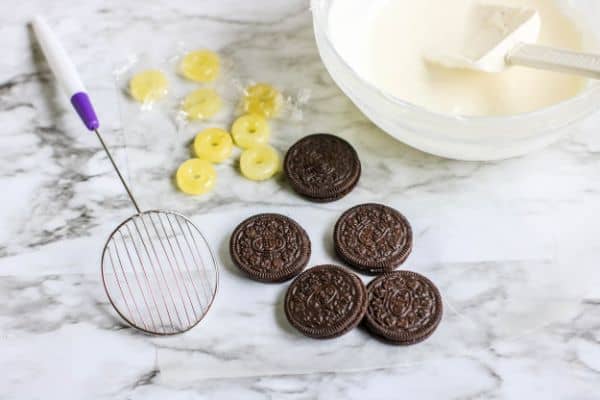 a bowl of melted white chocolate with a spatula in it next to yellow lifesavers. oreos and a strainer on a kitchen counter