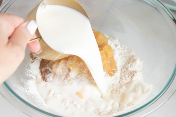 a hand pouring milk into the ingredients for apple cheesecake pancakes in a glass mixing bowl