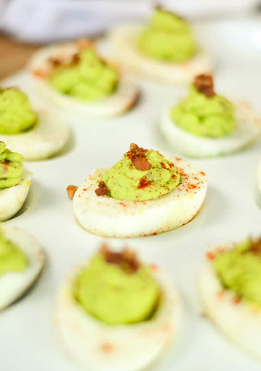 avocado and bacon deviled eggs on a white dish, one is seen clearly, the rest are blurry