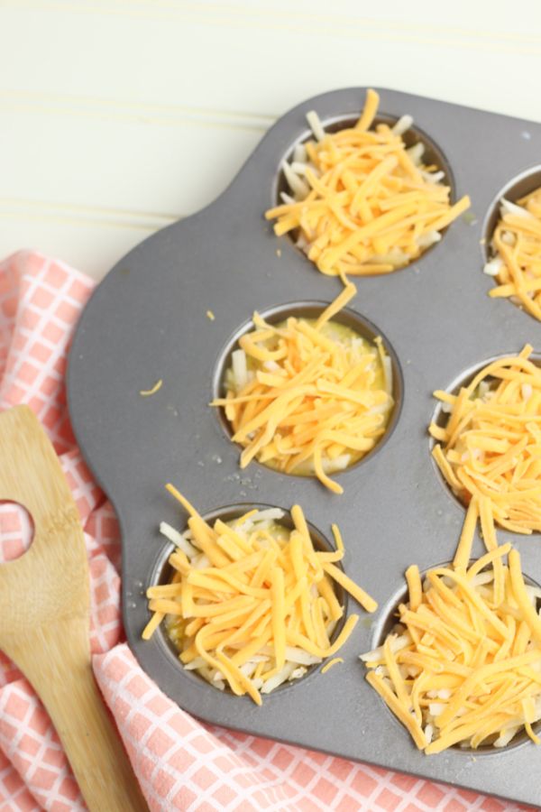 bacon and egg cups in a muffin tin before baking next to a peach colored linen next to a wooden spoon