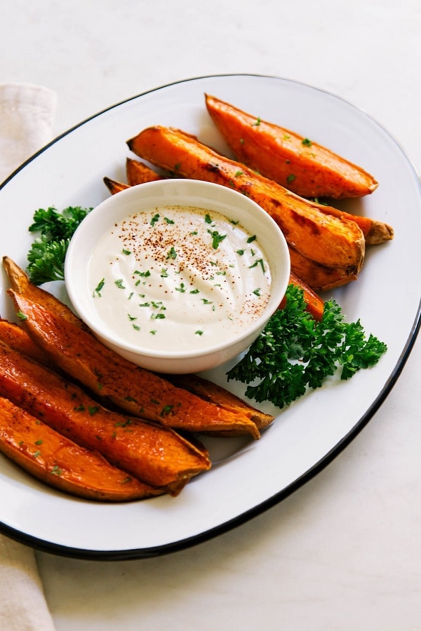 baked sweet potato wedges on a white dish with a bowl of dipping sauce and parsley on a white table