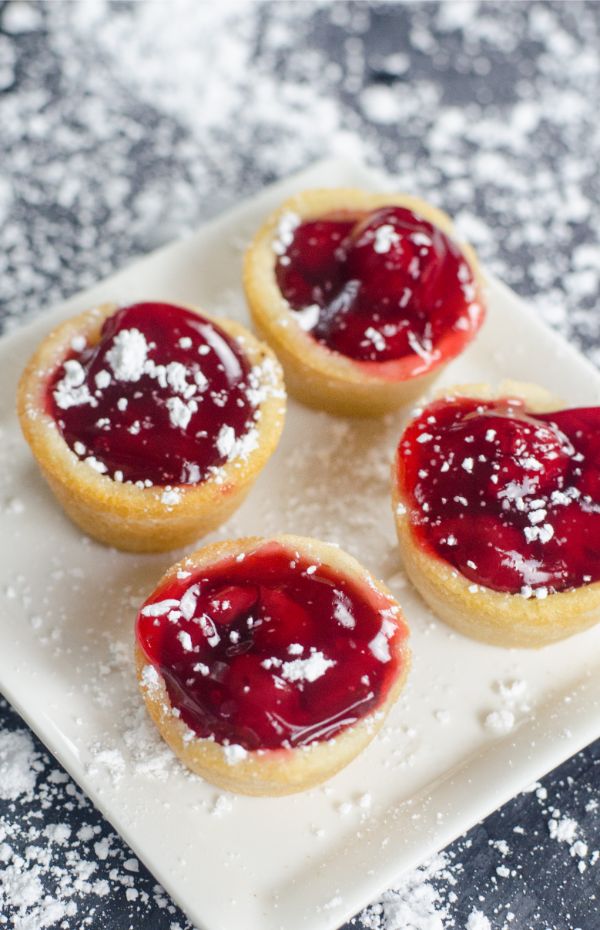 cherry pie filling in cookie cups sprinkled with powder sugar on a white plate on a gray counter with powder sugar on it