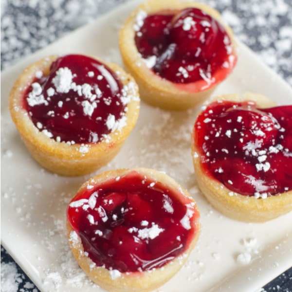 cherry pie filling in cookie cups sprinkled with powder sugar on a white plate on a gray counter with powder sugar on it