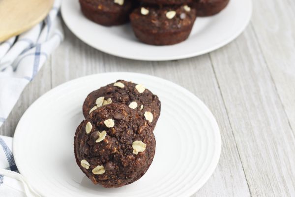 two Chocolate Banana Oat Chocolate Chip Muffins on a white plate on a table with another plate of muffins in the background