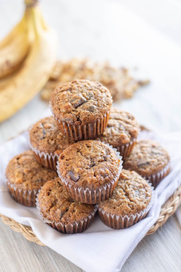 a stack of chunky monkey muffins on a white cloth on a brown bamboo plate on a brown table with bananas and walnuts in the background