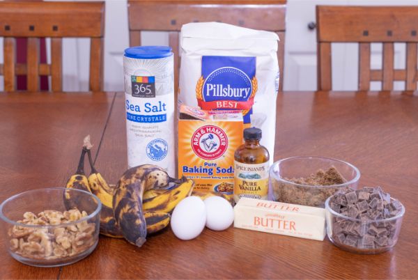 ingredients for chunky monkey muffins, glass bowls of walnuts, brown sugar and chocolate chunks, ripe bananas, two eggs, butter, containers of sea salt, baking soda, flour and vanilla, all on a brown table with chairs in the background