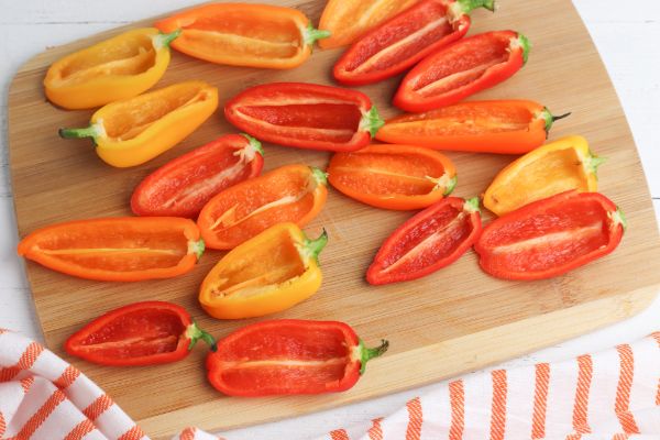 mini red and orange peppers halved, with the seeds removed on a wooden cutting board on a white wood table with an orange and white striped linen in the background