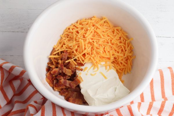 cream cheese, cheddar cheese, mayonnaise and bacon in a white bowl with an orange and white striped linen in the background on a white wood table
