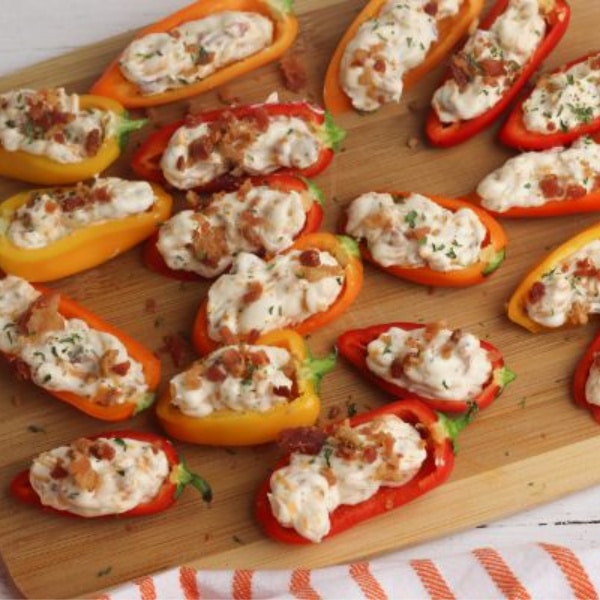 mini red and orange peppers stuffed with cream cheese, cheddar cheese, mayonnaise and bacon on a wooden cutting board on a white wood table with three stacked white plates and an orange and white striped linen in the background