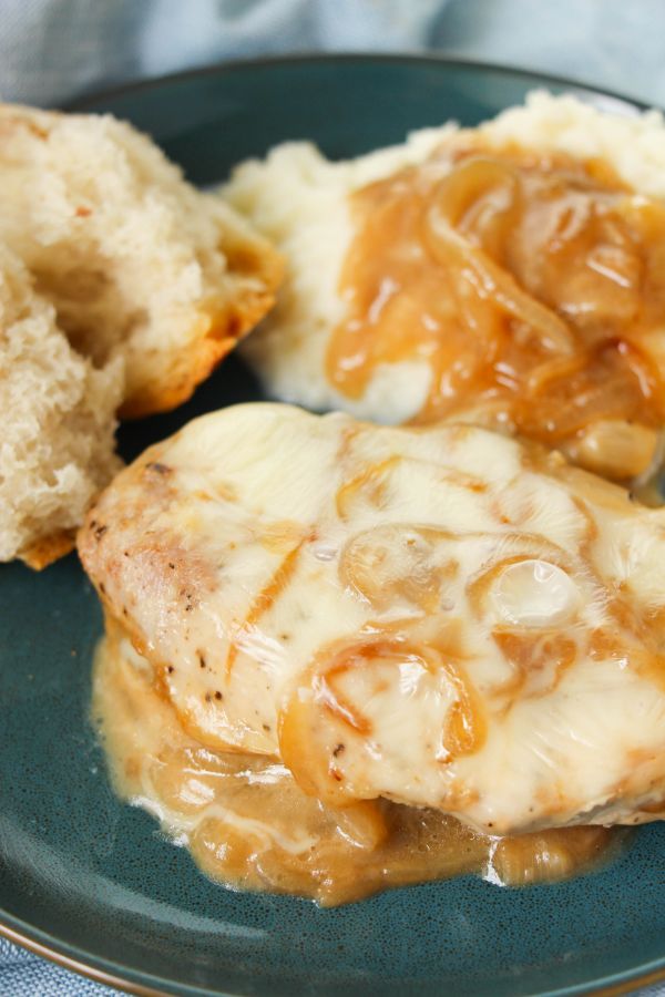 pork chops topped with sauce, cheese and onions on a plate next to mashed potatoes topped with onion and sauce, and bread
