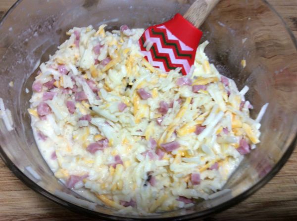 eggs, shredded cheese, diced ham, and hashbrowns being mixed together with a spatula in a glass bowl on a brown table